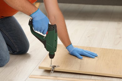 Photo of Man with electric screwdriver assembling furniture on floor indoors, closeup