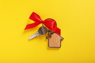 Key with trinket in shape of house and red bow on yellow background, top view. Housewarming party