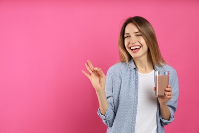 Photo of Young woman with glass of chocolate milk showing Ok on pink background. Space for text
