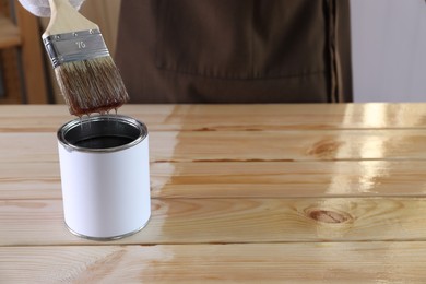 Man dipping brush into can with varnish at wooden table indoors, closeup