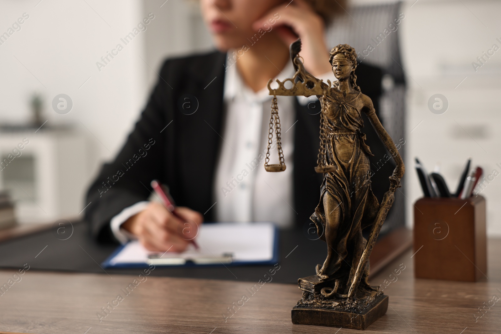 Photo of Notary with clipboard writing notes at workplace in office, focus on statue of Lady Justice