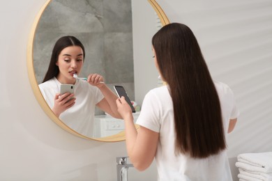 Photo of Beautiful young woman using smartphone while brushing teeth in bathroom. Internet addiction