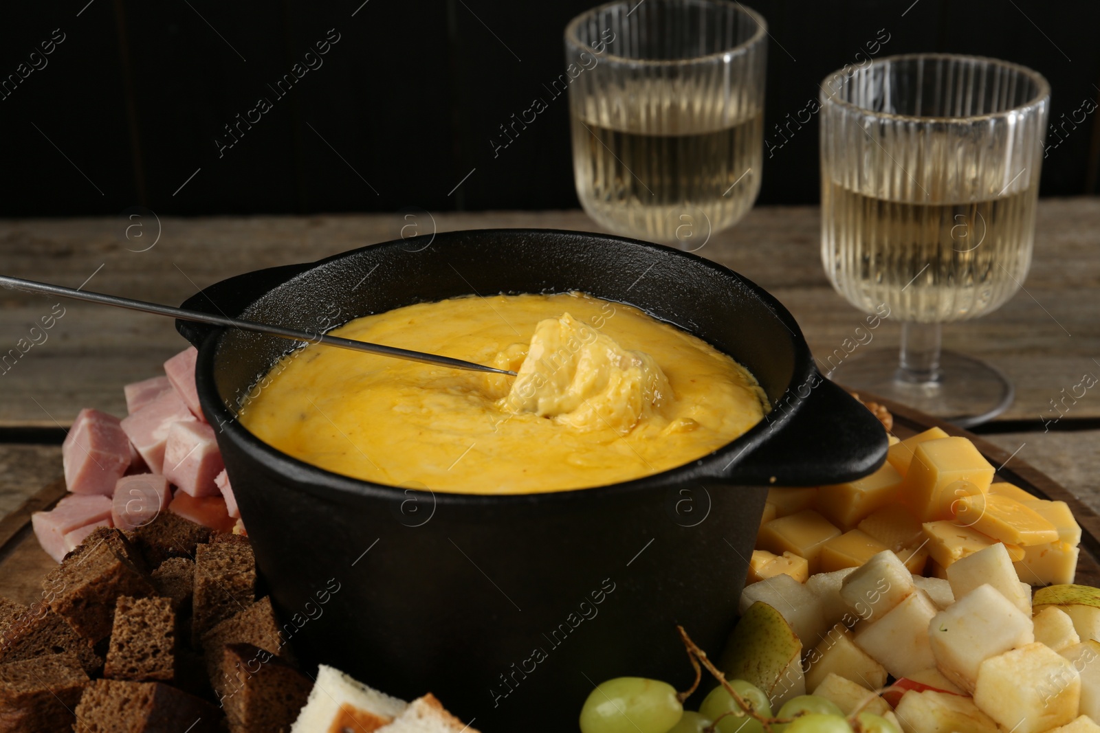 Photo of Fondue pot with melted cheese, glasses of wine and different products on wooden table, closeup