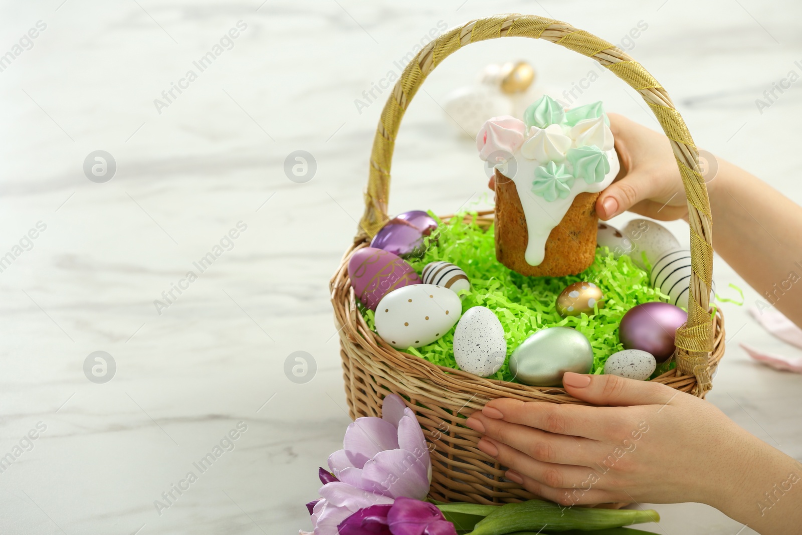 Photo of Woman putting delicious Easter cake with meringues into wicker basket at white marble table, closeup. Space for text