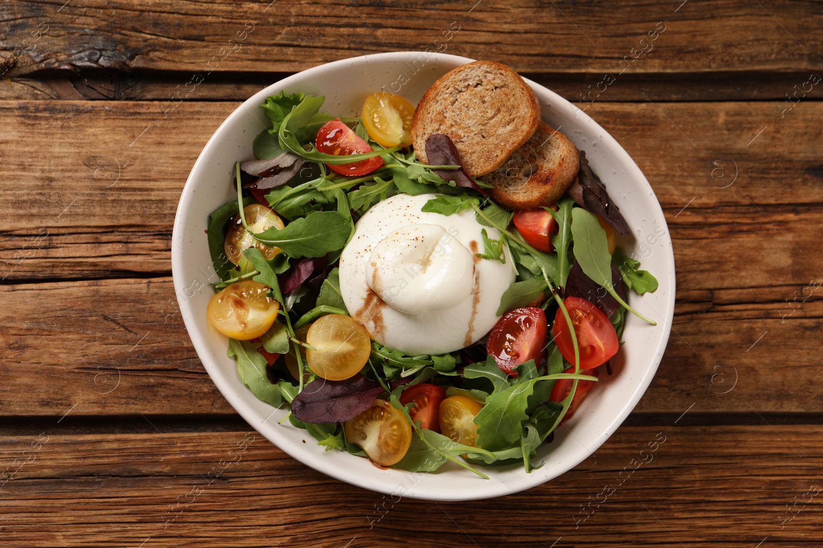 Photo of Delicious burrata salad with colorful cherry tomatoes, croutons and arugula on wooden table, top view