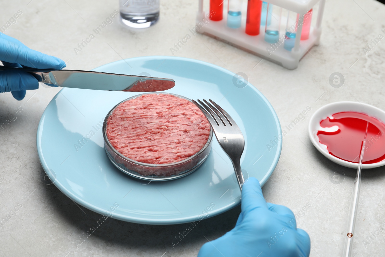 Photo of Scientist holding fork and knife over plate with minced cultured meat at light grey lab table, closeup