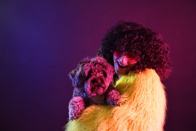 Beautiful young woman in yellow fur coat and sunglasses with cute dog on color background in neon lights. Space for text