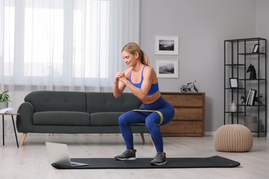 Photo of Fit woman doing squats with fitness elastic band near laptop on mat at home