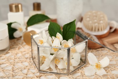 Glass box with beautiful jasmine flowers and skin care products on table, closeup