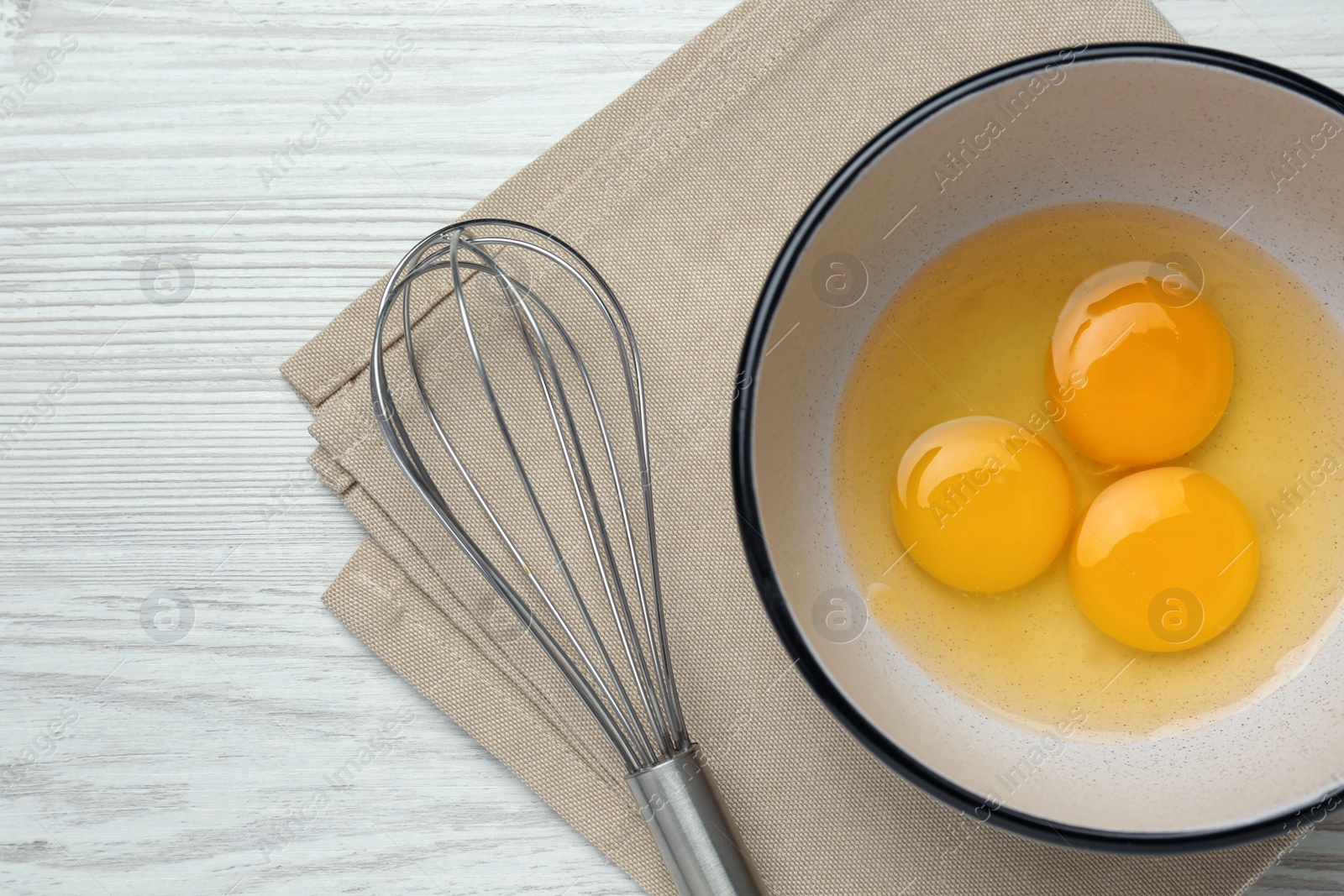 Photo of Whisk near bowl with eggs on white wooden table, top view. Space for text