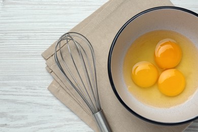 Photo of Whisk near bowl with eggs on white wooden table, top view. Space for text