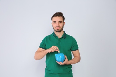 Young man putting money into piggy bank on light background