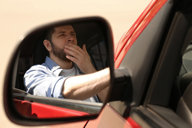 Photo of Tired man yawning in his auto, view through car side mirror