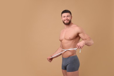 Photo of Smiling athletic man measuring waist with tape on brown background, space for text. Weight loss concept