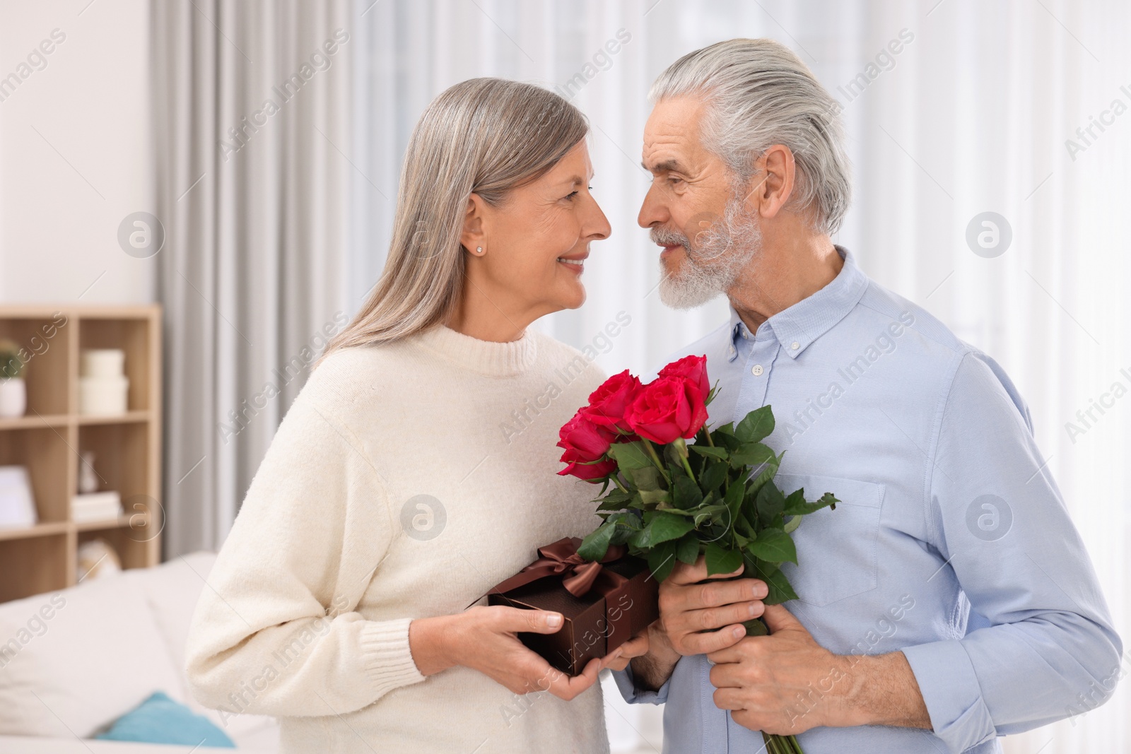 Photo of Happy affectionate senior couple with gift box and beautiful bouquet indoors