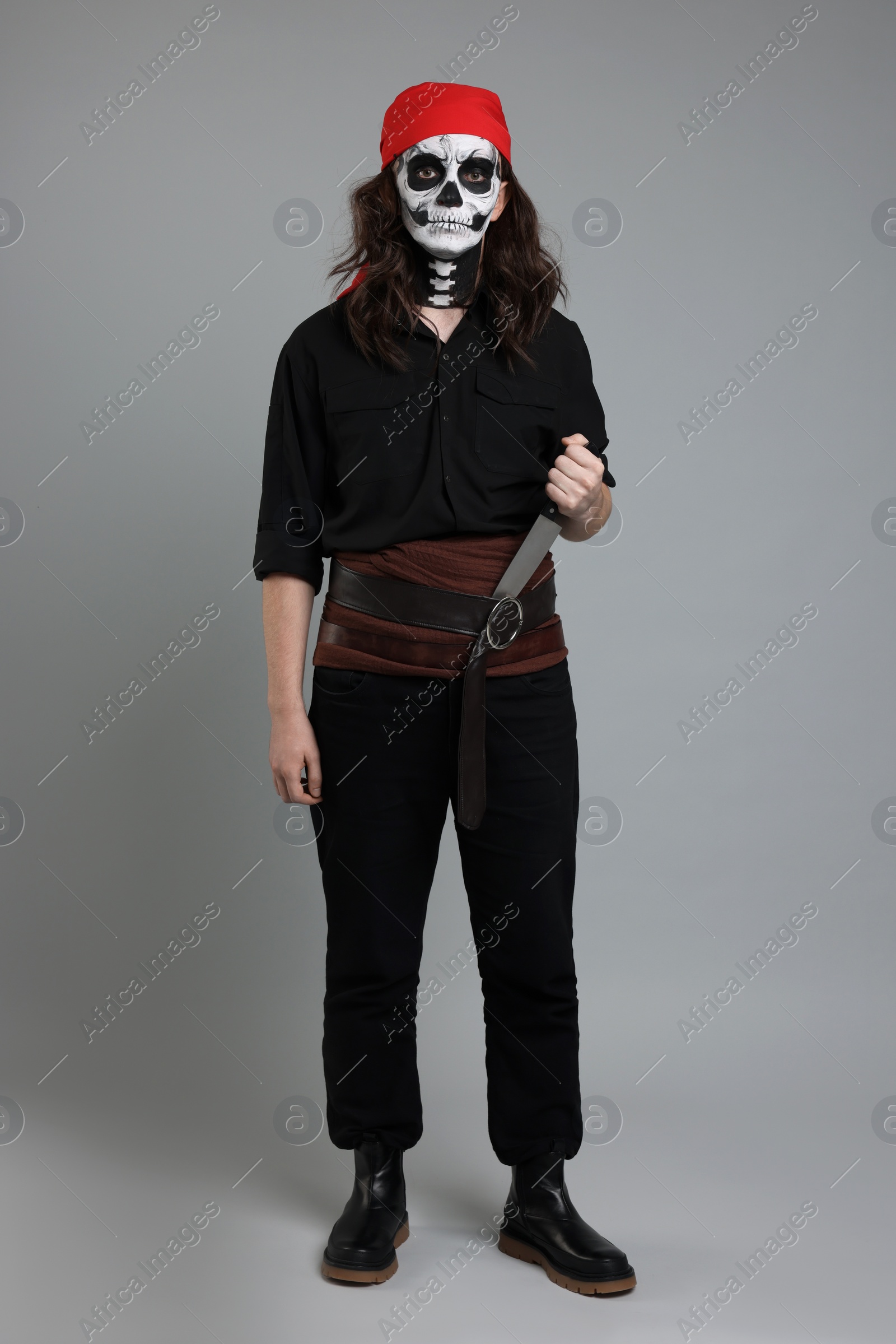 Photo of Man in scary pirate costume with skull makeup and knife on light grey background. Halloween celebration