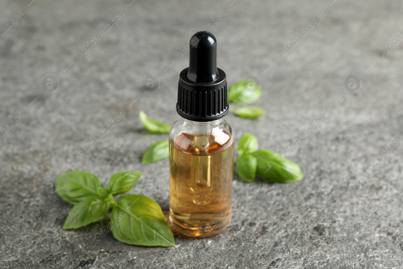 Photo of Glass bottle of basil essential oil and leaves on grey stone table