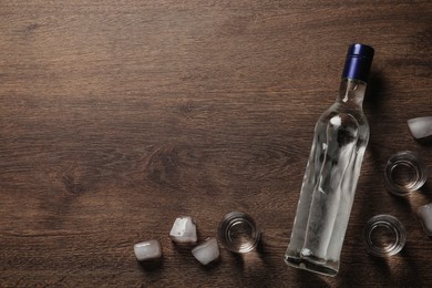 Photo of Bottle of vodka, shot glasses and ice cubes on wooden table, flat lay. Space for text