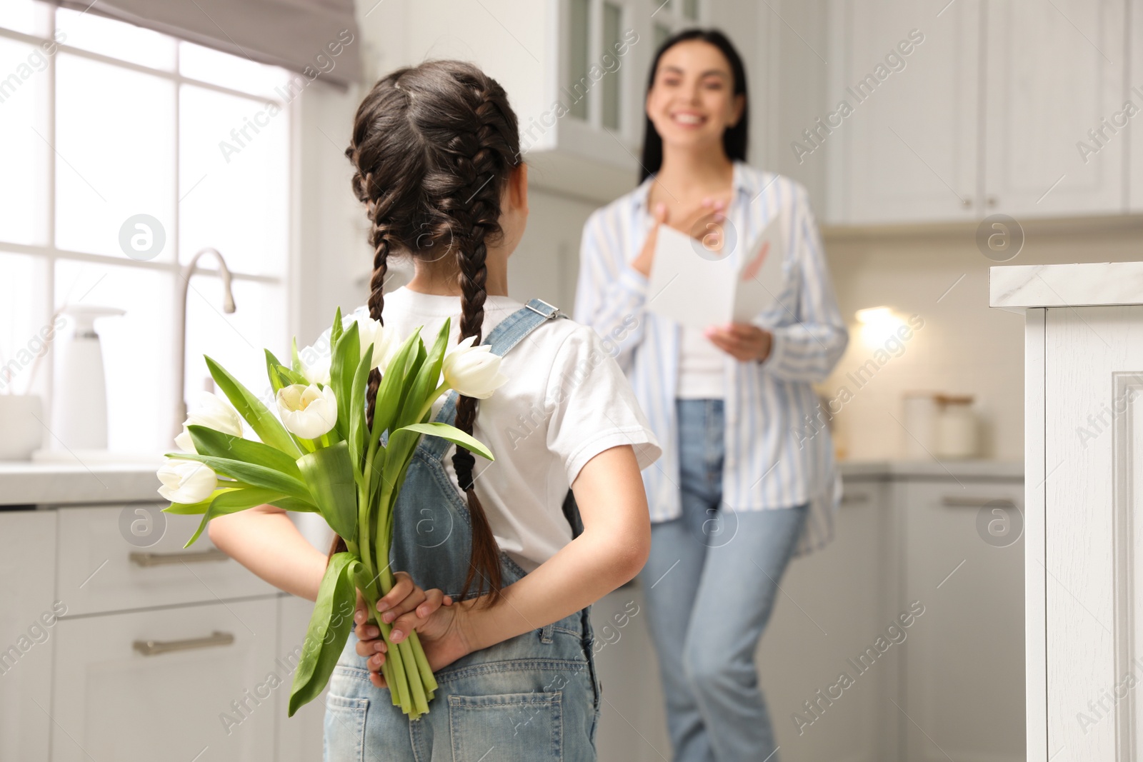 Photo of Little girl hiding tulip bouquet for mom in kitchen at home. Happy Mother's Day