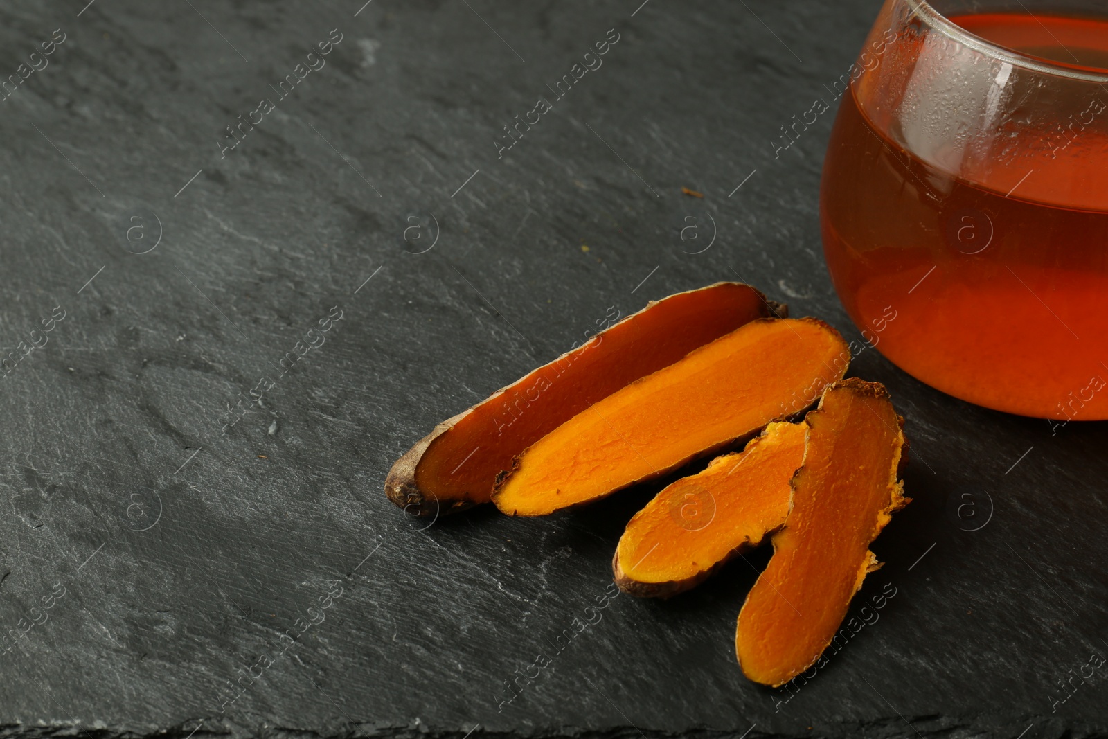 Photo of Glass cup of tasty tea and cut turmeric roots on black textured table, space for text