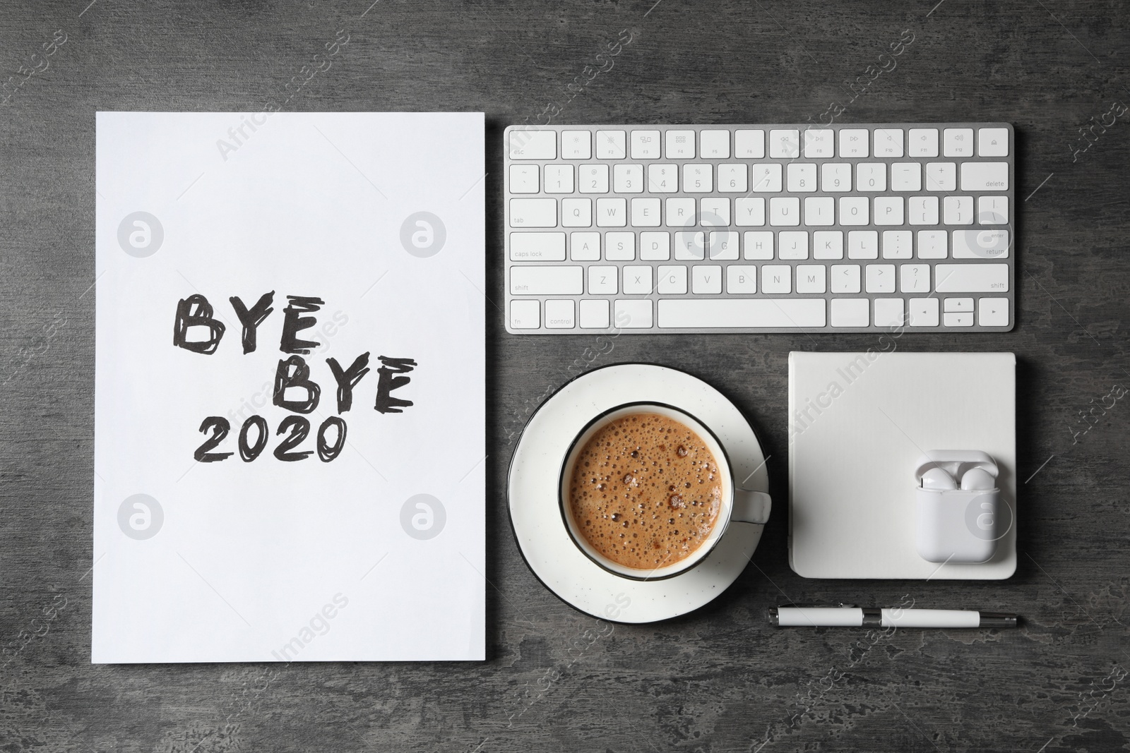 Photo of White sheet of paper with phrase Bye Bye 2020 near coffee, keyboard and wireless earphones on grey table, flat lay