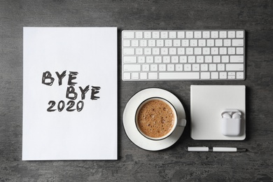 Photo of White sheet of paper with phrase Bye Bye 2020 near coffee, keyboard and wireless earphones on grey table, flat lay