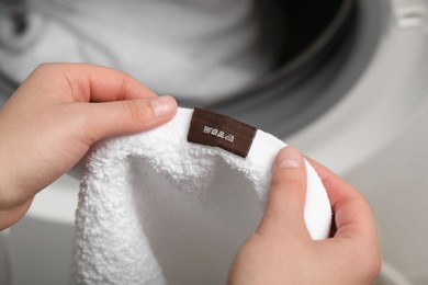 Woman reading clothing label with care symbols and material content on white towel near washing machine, closeup