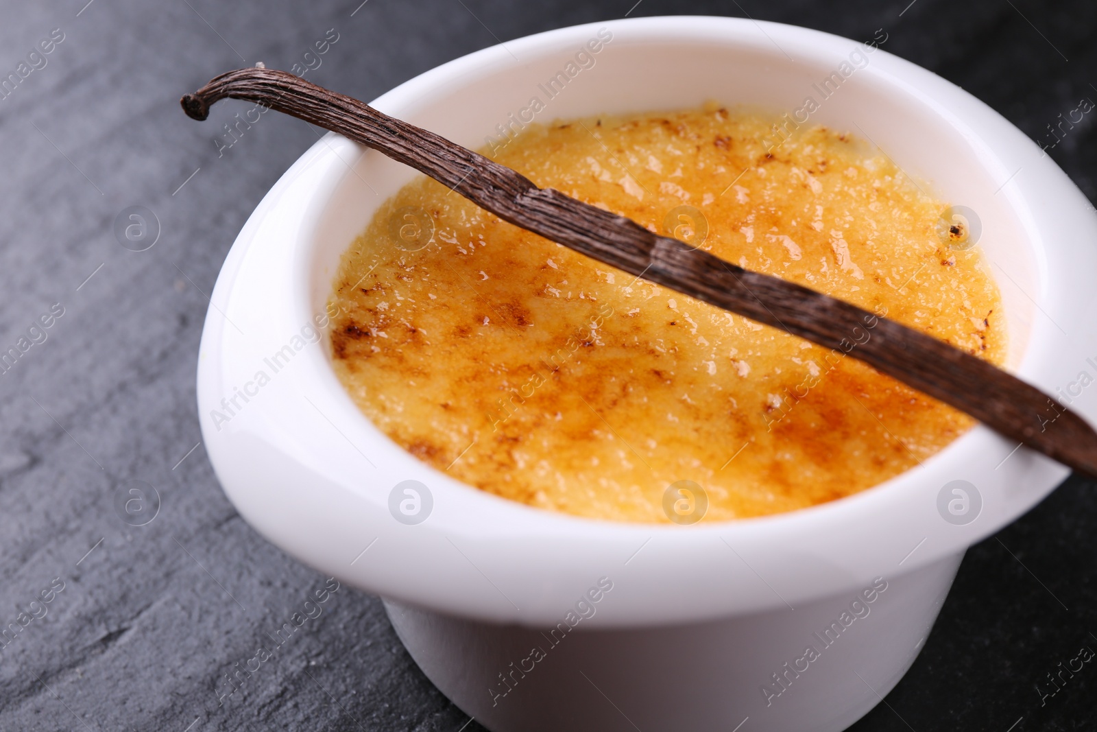 Photo of Delicious creme brulee in bowl and vanilla pod on dark gray textured table