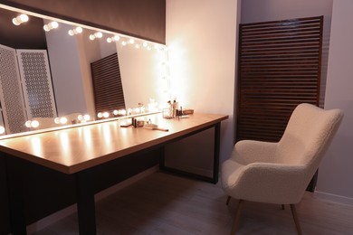 Stylish mirror with light bulbs near table with makeup products and armchair in dressing room