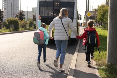 Photo of Being late for school. Senior woman and her grandchildren with backpacks running towards bus outdoors, back view