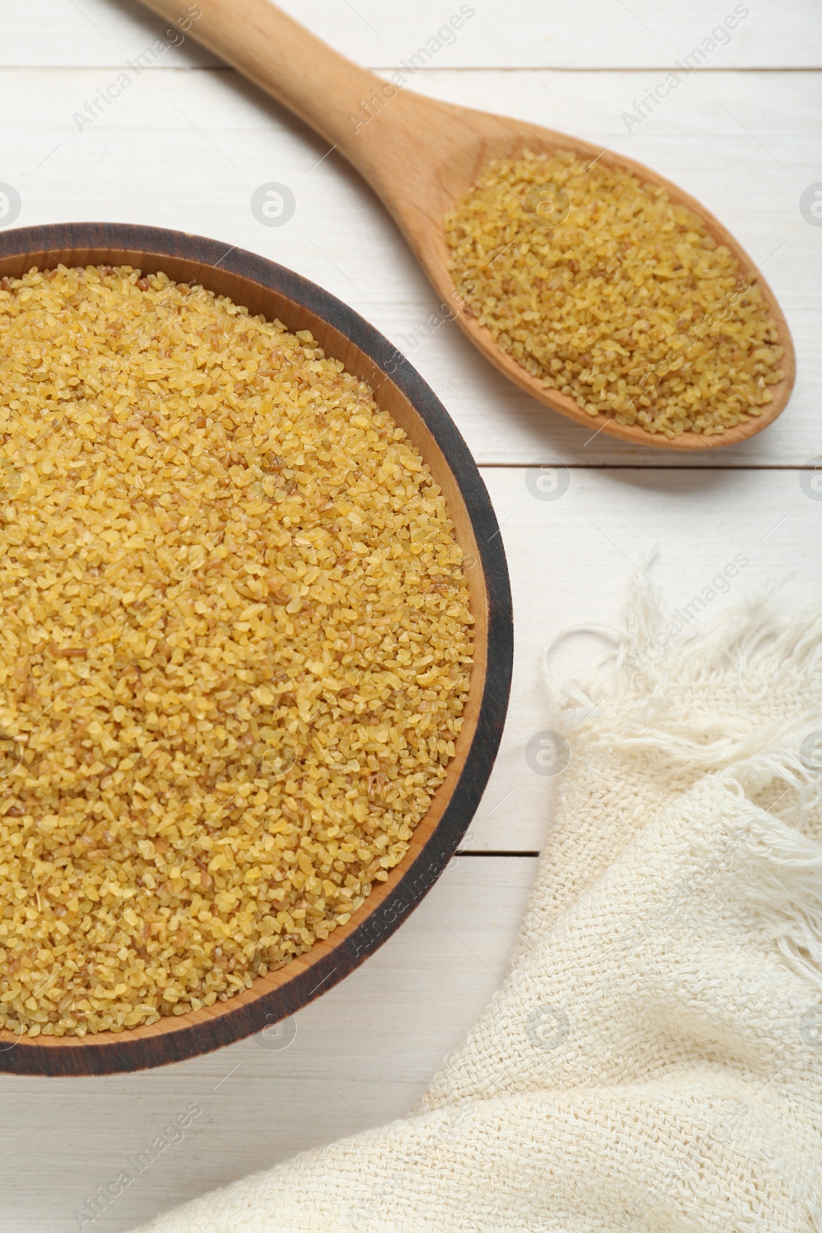 Photo of Bowl and spoon with uncooked bulgur on white wooden table, flat lay