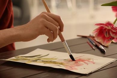 Photo of Woman painting flowers with watercolor at wooden table indoors, closeup. Creative artwork