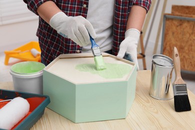 Photo of Man painting honeycomb shaped shelf with brush at wooden table indoors, closeup