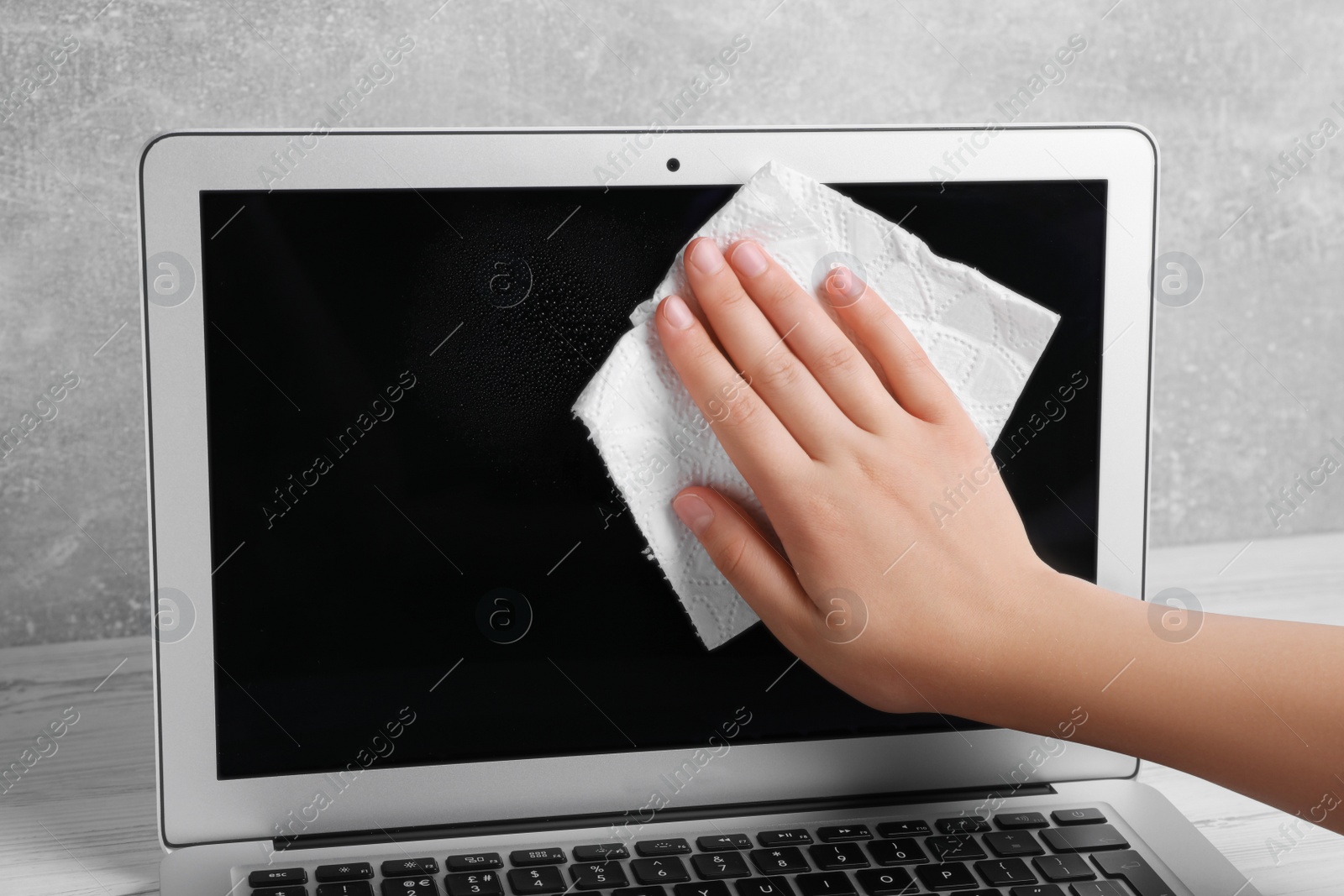 Photo of Woman wiping laptop with paper towel at white wooden table, closeup