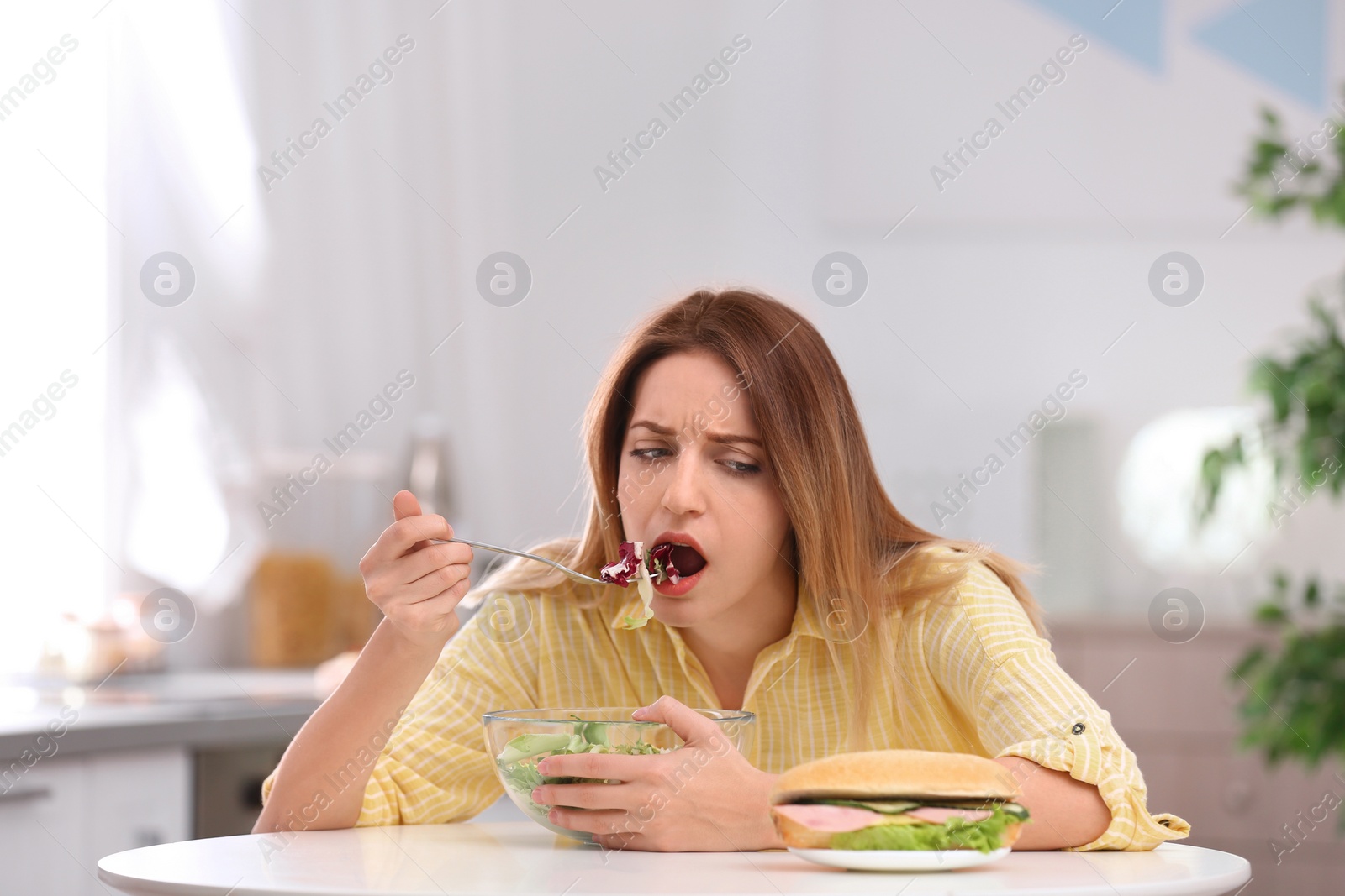 Photo of Emotional young woman eating salad instead of sandwich in kitchen. Healthy diet