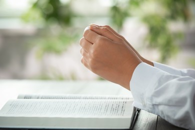 Woman holding hands clasped while praying at grey table with Bible, closeup