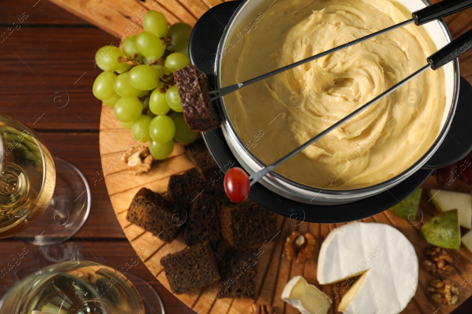 Photo of Forks with pieces of grape, bread, fondue pot with melted cheese, wine and snacks on wooden table, flat lay