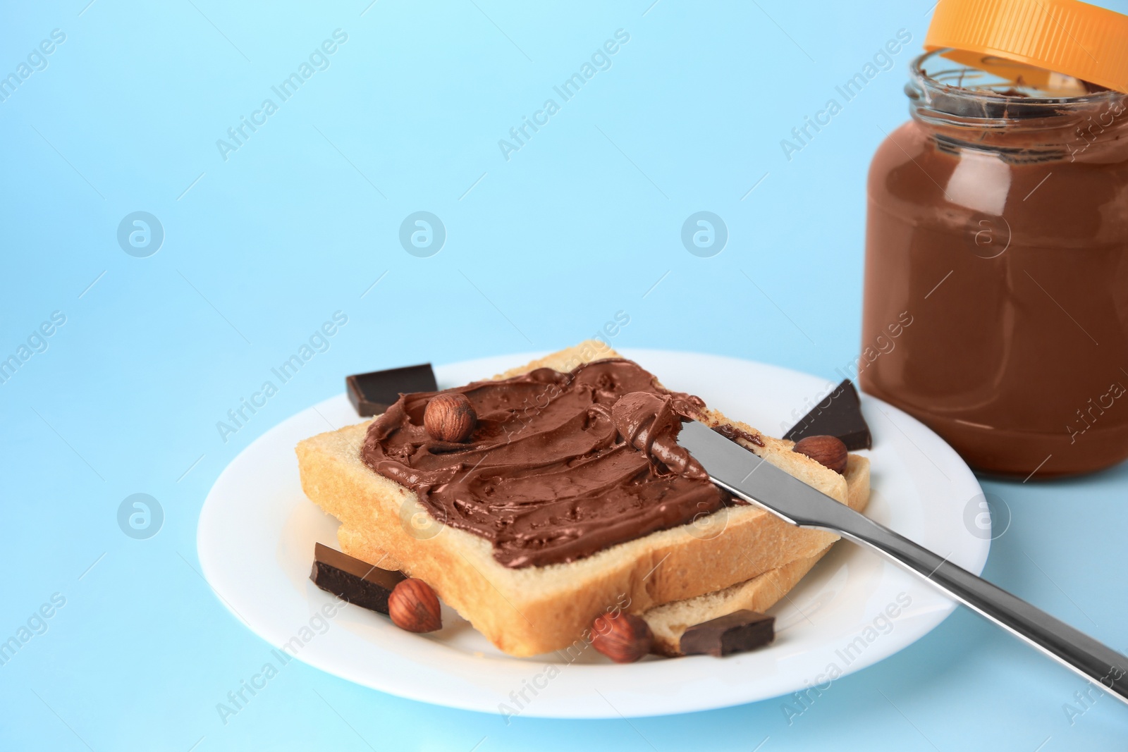 Photo of Tasty toast with chocolate paste and nuts near jar of cream on light blue background