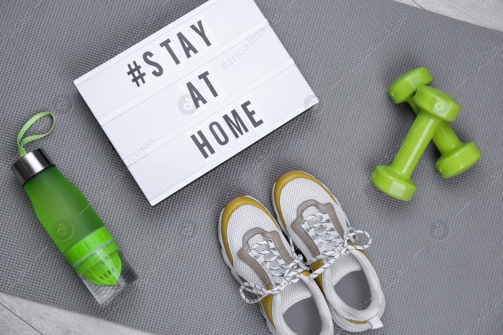 Photo of Sport equipment and lightbox with hashtag STAY AT HOME on grey yoga mat, flat lay. Message to promote self-isolation during COVID‑19 pandemic