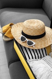 Photo of Open suitcase full of clothes, shoes and summer accessories on sofa
