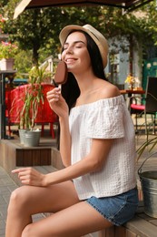 Photo of Beautiful young woman eating ice cream glazed in chocolate on city street
