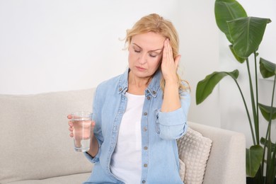 Photo of Woman with glass of water suffering from headache on sofa at home. Hormonal disorders