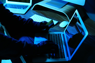 Hacker with credit card and laptop in dark room, closeup. Cyber crime