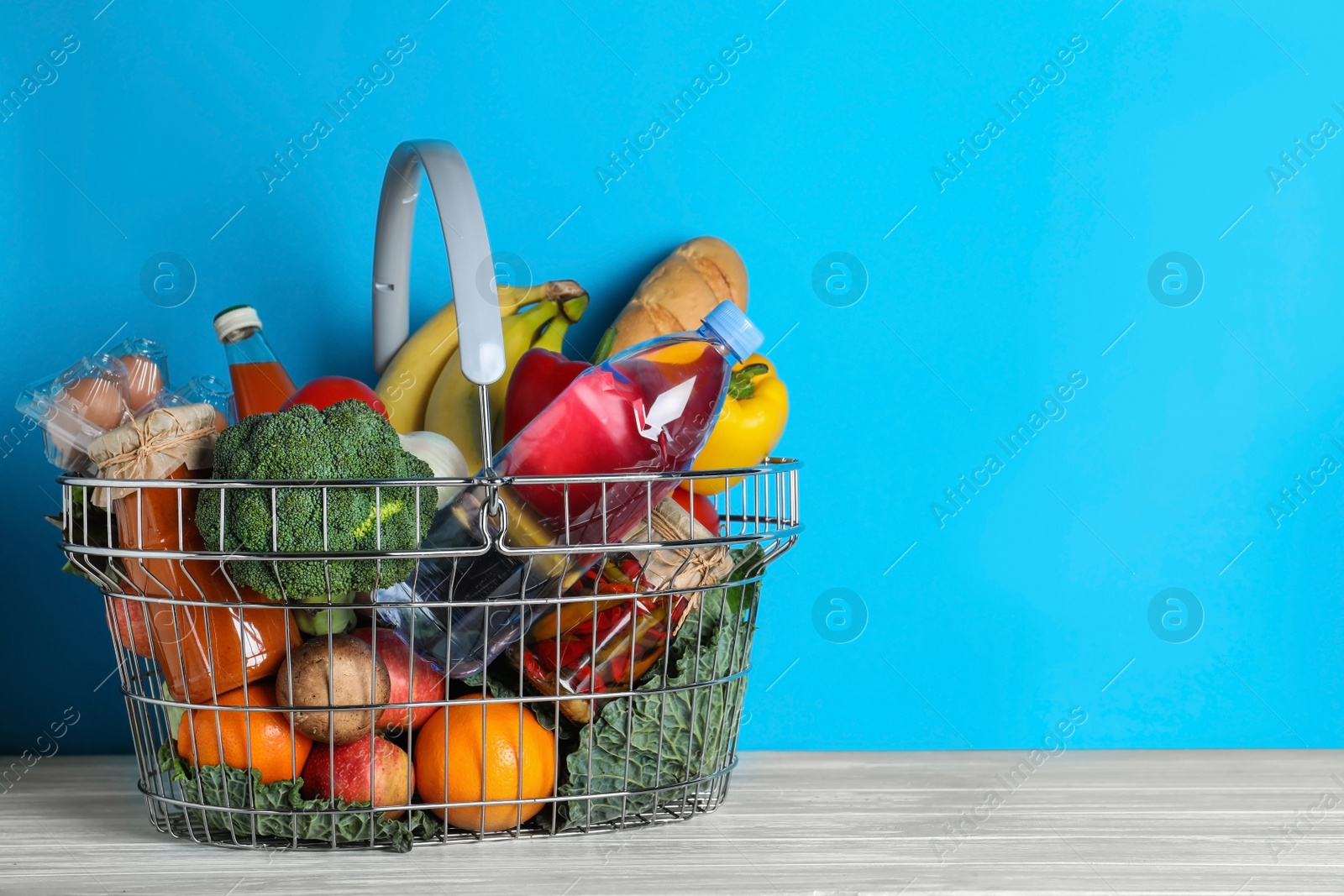 Photo of Shopping basket with grocery products on white wooden table against  light blue background. Space for text