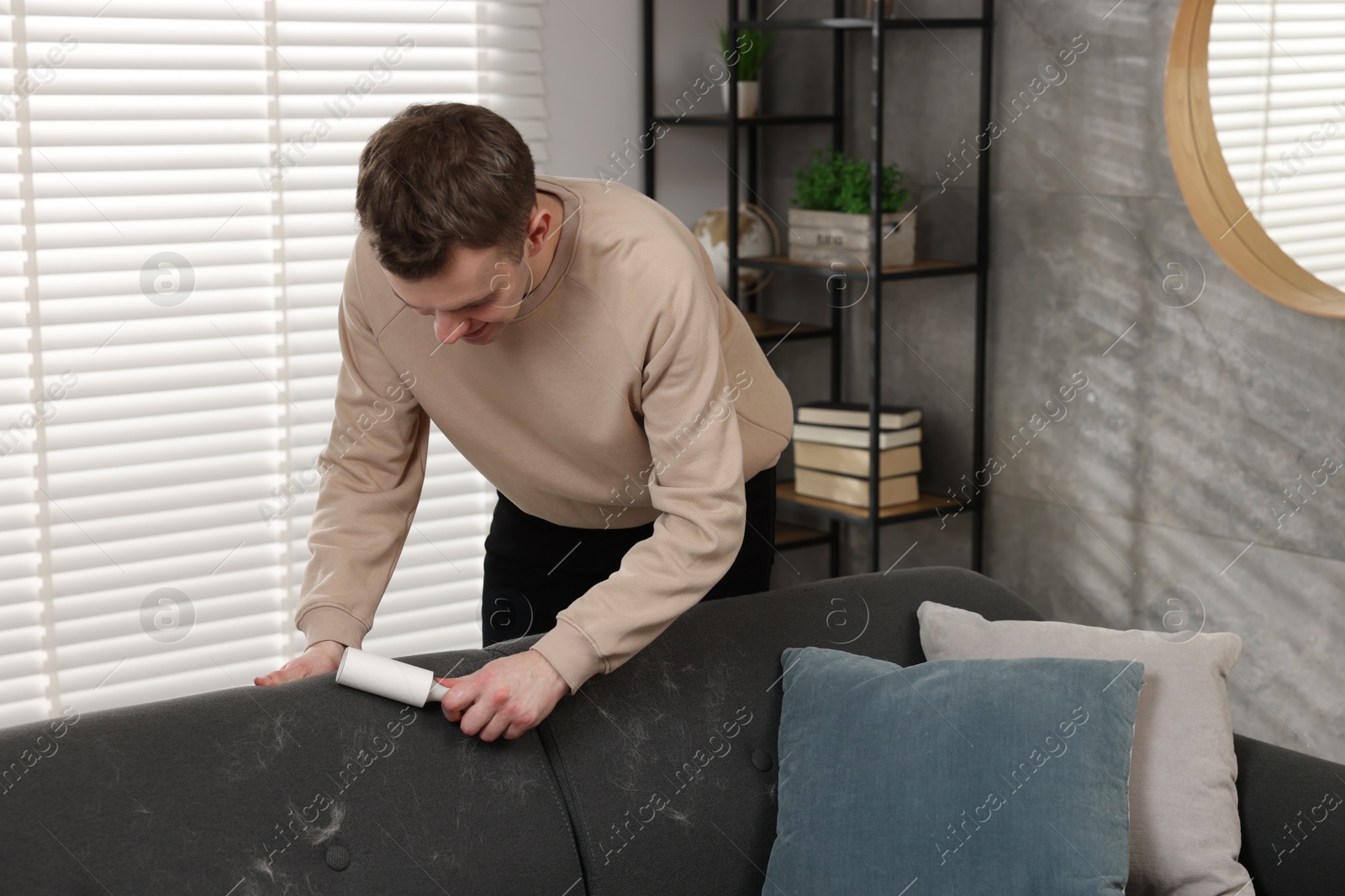 Photo of Pet shedding. Smiling man with lint roller removing dog's hair from sofa at home