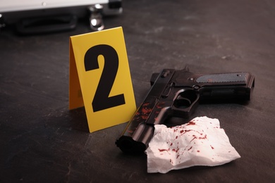 Napkin with blood, gun and evidence marker on black slate table, closeup. Crime scene