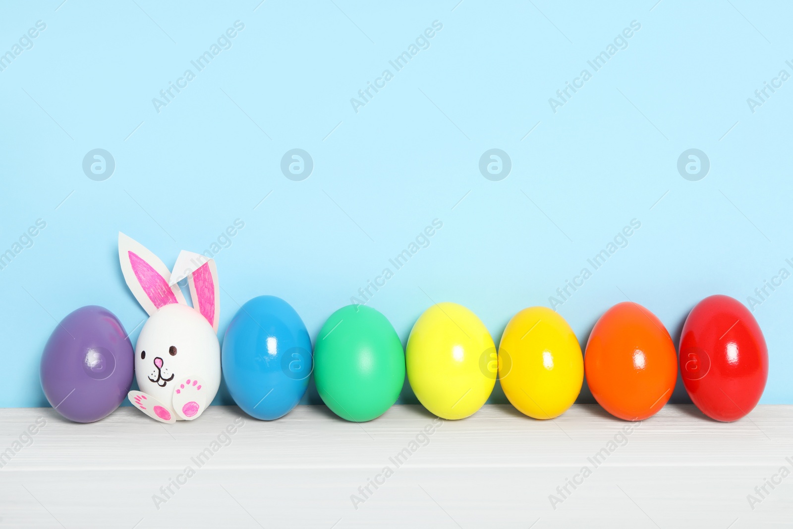 Photo of Bright Easter eggs and white one as cute bunny on wooden table against light blue background, space for text