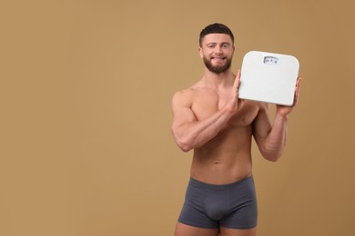 Photo of Smiling athletic man holding scales on brown background, space for text. Weight loss concept