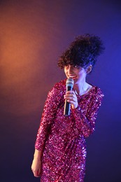 Beautiful young woman with microphone singing on color background in neon lights