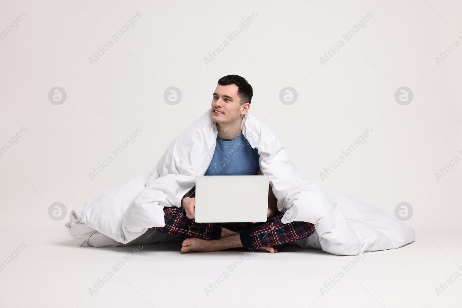 Photo of Happy man in pyjama wrapped in blanket using laptop on light grey background